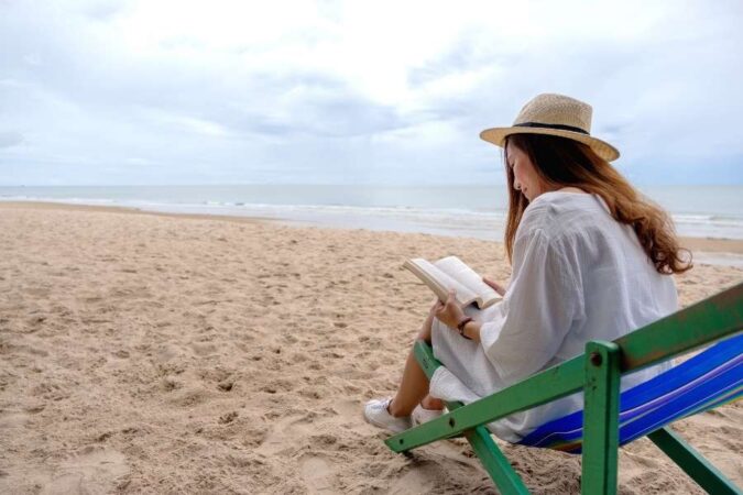 Une femme portant un chapeau de paille profite d'un moment de lecture et détente assise dans une chaise longue sur la plage.